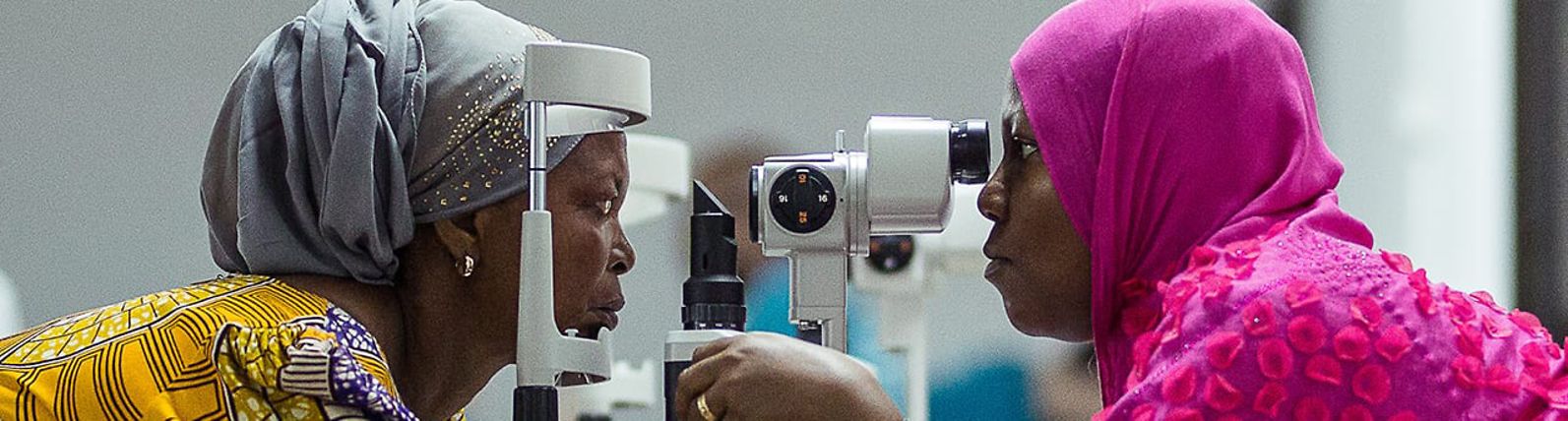 A woman examines another woman's eyes using a slit lamp.